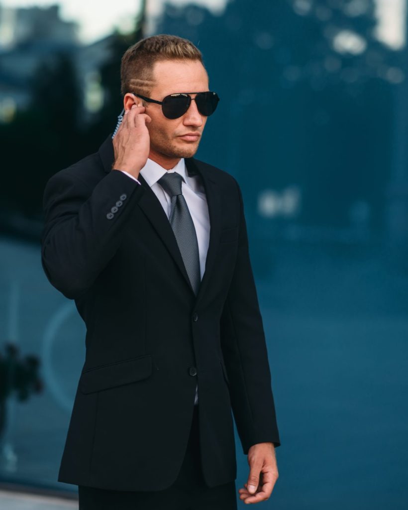 Male bodyguard uses security earpiece outdoors, professional communication tools. Guarding is a risky profession. Guard in suit and sunglasses